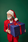 cheerful man dressed as Santa holding huge present smiling and looking away, Christmas concept magic mug #681098286