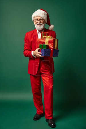 elegante Papá Noel en rojo elegante atuendo celebración de la pila de regalos sobre fondo verde, concepto de invierno