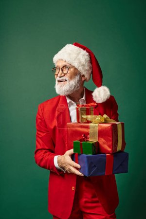 cheerful Santa in christmassy hat holding pile of presents smiling looking away, winter concept