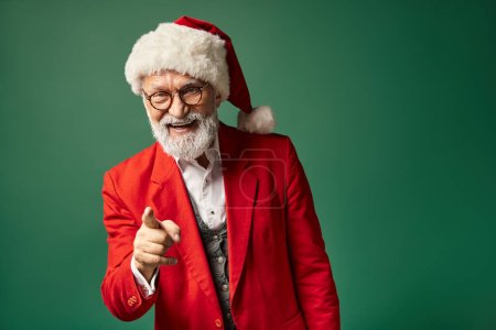 joyful elegant Santa with beard smiling cheerfully and pointing finger at camera, winter concept
