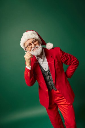 fröhlich eleganter Mann als Weihnachtsmann mit Brille und Hut gekleidet posiert vor dunkelgrünem Hintergrund, Winterkonzept
