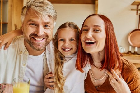 excited parents with cute daughter looking at camera in kitchen at home, family interaction