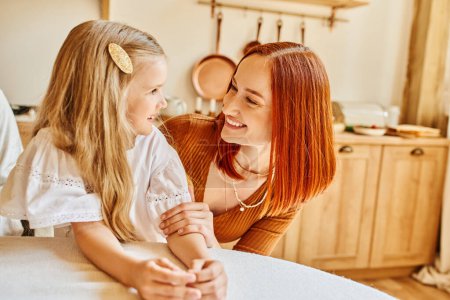cheerful redhead woman and cute daughter looking at each other in cozy kitchen, love and care