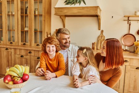 smiling parents with daughter and son sitting near fresh fruits and orange juice in cozy kitchen
