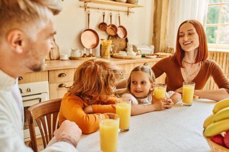 Foto de Padres alegres con hija e hijo cerca de zumo de naranja y frutas durante el desayuno en la cocina - Imagen libre de derechos