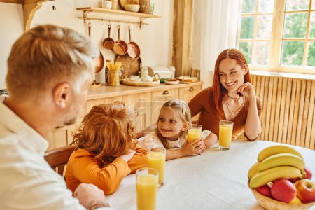 fröhliche Geschwister lächeln einander in der Nähe von frischem Orangensaft und Eltern in der heimischen Küche an