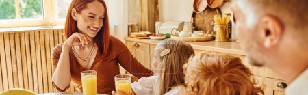 joyful woman looking at children near fresh orange juice during breakfast in cozy kitchen, banner