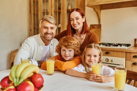 joyful couple with adorable kids looking at camera near fresh fruits and orange juice in kitchen