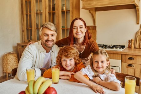 glückliche Eltern mit entzückenden Kindern, die in der Nähe von frischem Obst und Orangensaft in die Kamera schauen