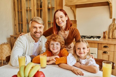 freudige Eltern mit entzückenden Kindern, die in der Küche neben frischem Obst und Orangensaft in die Kamera schauen