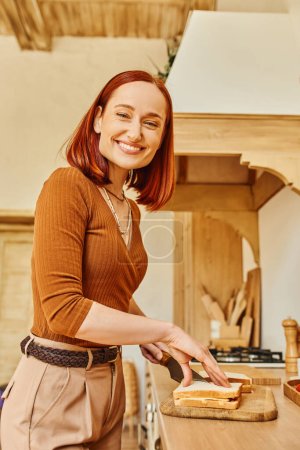 joyful redhead woman looking at camera and preparing delicious sandwiches for breakfast in kitchen