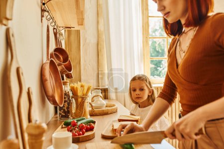 Téléchargez les photos : Mère souriante légumes coupe préparer le petit déjeuner près de la fille avec sandwich sur le comptoir de la cuisine - en image libre de droit