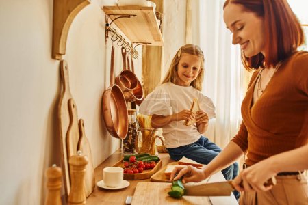 Foto de Mujer feliz cortando verduras preparando el desayuno cerca de la hija con sándwich en el mostrador de cocina - Imagen libre de derechos