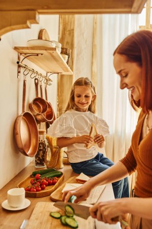 niño alegre sentado con sándwich en el mostrador de la cocina cerca de la madre sonriente preparando el desayuno