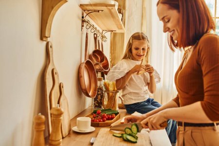 Foto de Feliz hija sentada con sándwich en el mostrador de la cocina cerca de la madre sonriente preparando el desayuno - Imagen libre de derechos
