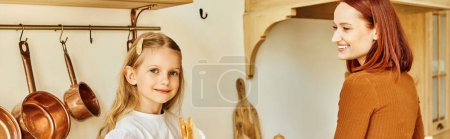 Photo for Cute girl with sandwich looking at camera near mother cooking breakfast in kitchen, banner - Royalty Free Image
