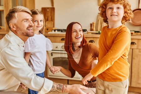fröhliche Eltern mit unbeschwerten Kindern, die sich beim Spielen in der Küche an den Händen halten, gehegte Erinnerungen