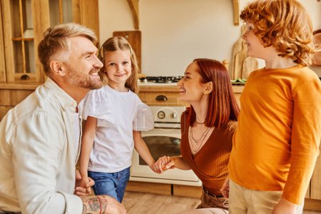 smiling parents with carefree kids holding hands while having fun in kitchen, emotional connection