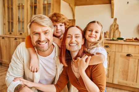 joyful parents with children embracing and looking at camera in cozy kitchen, cherished moments