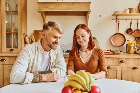 mujer sonriente mirando teléfono inteligente cerca de marido y frutas frescas mientras está sentado en la cocina moderna