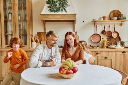 Photo for Playful kids running around happy parents sitting with smartphone near fresh fruits in cozy kitchen - Royalty Free Image