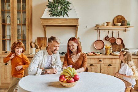 joyful kids running around happy parents sitting with smartphone near fresh fruits in cozy kitchen