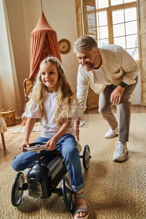 Photo for Happy man assisting cheerful daughter riding toy car in modern living room at home, playing together - Royalty Free Image