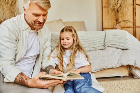 Téléchargez les photos : Père attentionné et fille mignonne assis sur le sol dans la chambre et le livre de lecture, apprendre ensemble - en image libre de droit