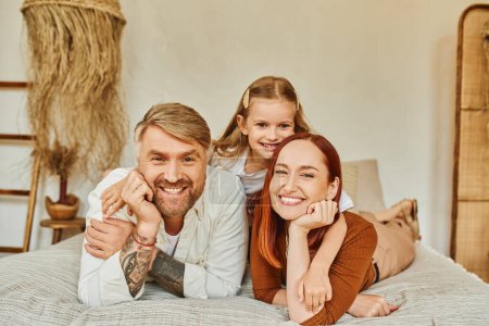 Photo for Cheerful parents with carefree daughter relaxing on bed and looking at camera, cozy home environment - Royalty Free Image