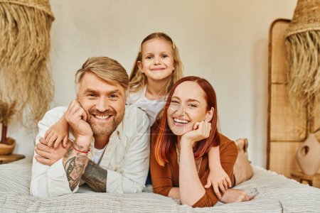 Photo for Happy tattooed man with smiling wife and adorable daughter looking at camera on bed at home - Royalty Free Image