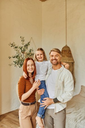 Photo for Cheerful husband and wife holding cute daughter and looking at camera in cozy bedroom, love and care - Royalty Free Image