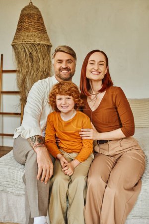 freudige Eltern mit rothaarigem Sohn, der im gemütlichen häuslichen Umfeld auf dem Bett sitzt und in die Kamera blickt