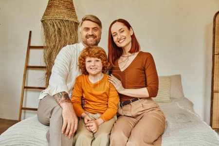 Photo for Cheerful parents with redhead son sitting on bed and looking at camera in cozy home environment - Royalty Free Image