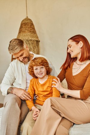smiling husband and wife embracing redhead kid while sitting in bedroom, emotional connection
