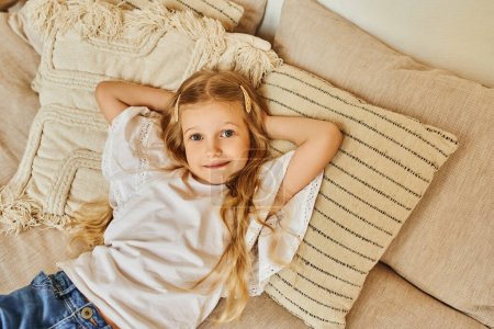 glückliches blondes Mädchen liegt auf weichen Kissen mit den Händen hinter dem Kopf und blickt in die Kamera im gemütlichen Schlafzimmer