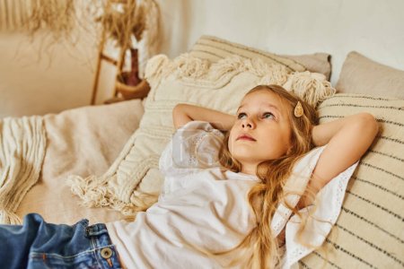 charming and dreamy girl lying on pillows with hands behind head and looking away in cozy bedroom