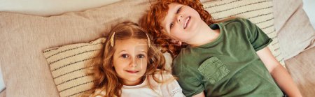 top view of siblings lying on bed with soft pillows and looking at camera in cozy bedroom, banner