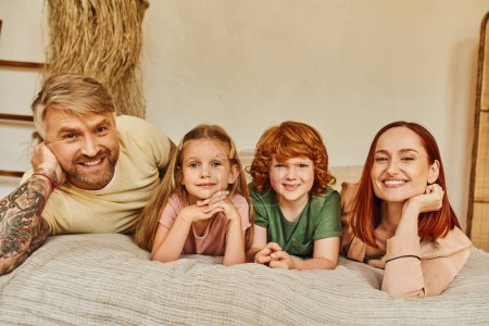 Lächelnde Eltern mit Sohn und Tochter im gemütlichen Schlafzimmer liegend und in die Kamera blickend, verbindende Momente