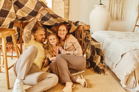 cheerful parents with happy daughter hiding under blanket hut in living room, playing together