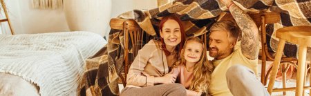 joyful parents with happy daughter playing under blanket hut in living room, horizontal banner