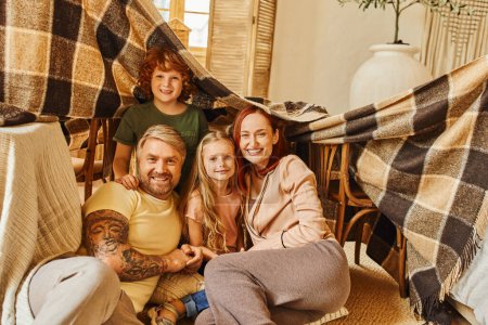 joyful parents and kids laughing under blanket hut in living room, playing together at home