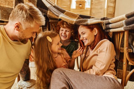 Aufgeregte Kinder mit lachenden Eltern unter Decke im Wohnzimmer, emotionale Verbindung