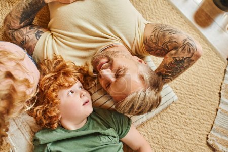 Photo for Top view of cheerful tattooed man lying down with redhead son on floor in living room, bonding - Royalty Free Image