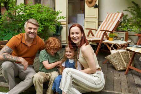 Foto de Padres riendo con niños felices sentados cerca de casa remolque y mirando a la cámara, tiempo en familia - Imagen libre de derechos