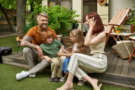 joyful couple with adorable children sitting near trailer home outdoors, family recreation