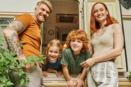 joyful couple looking at camera near kids having fun in trailer home, family leisure and recreation