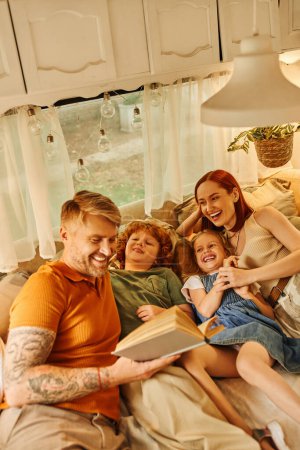 tattooed man reading book to laughing family on soft bed in cozy trailer home, learning together
