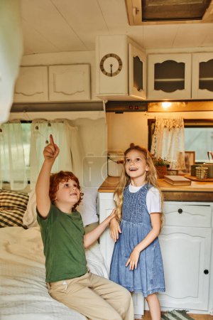 redhead boy pointing up in trailer home with modern cozy interior near amazed sister, happy siblings