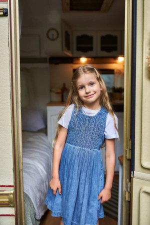 pleased girl with long blonde hair in stylish denim dress looking at camera from trailer home