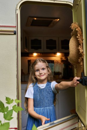 blonde girl in denim dress opening window on trailer home and looking at camera, happy childhood